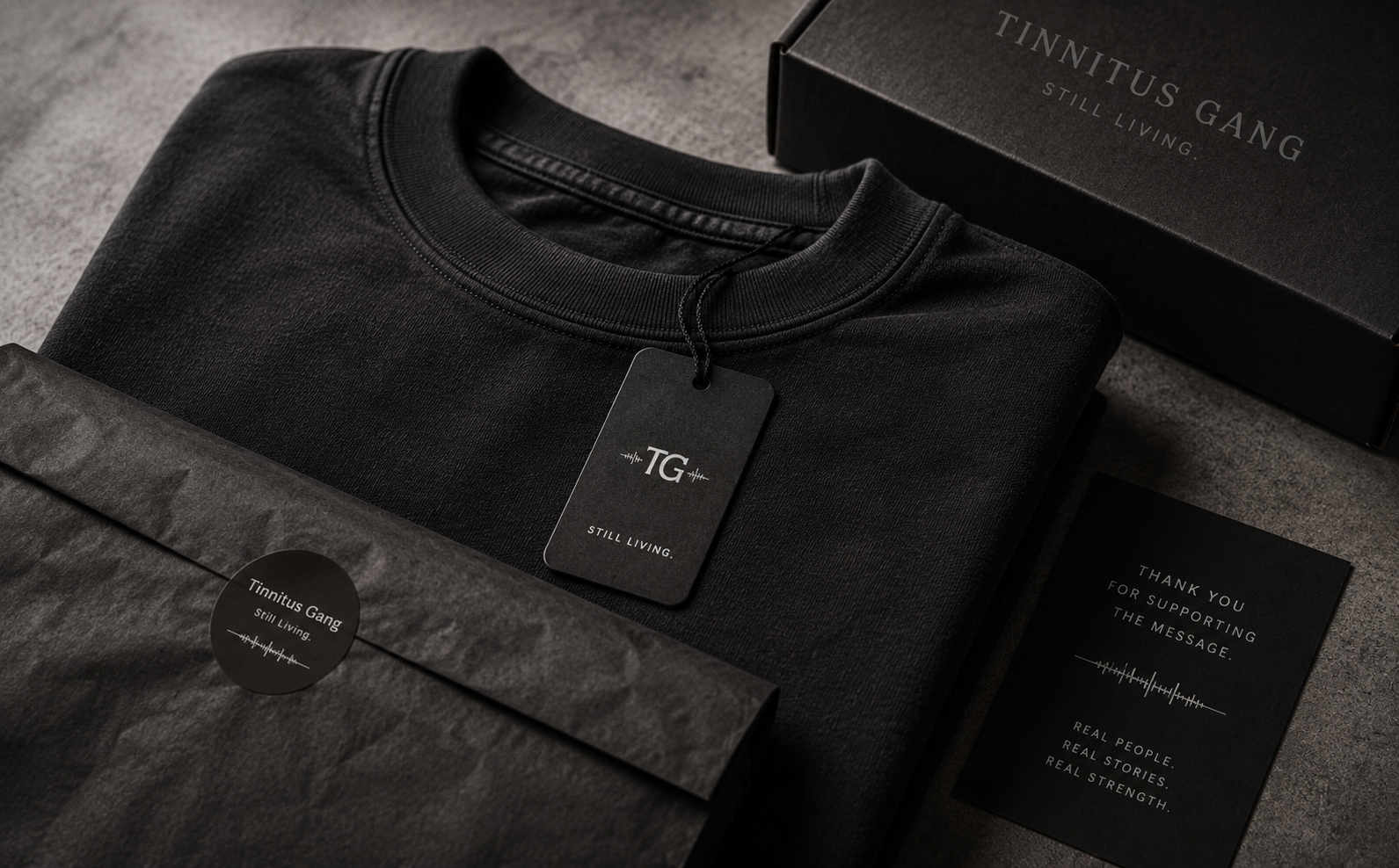 Folded black Tinnitus Gang tee with TG hangtag beside a matte black shipping box and thank-you card