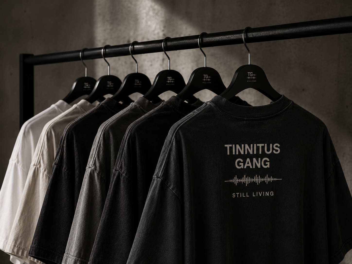 Rack of oversized Tinnitus Gang t-shirts in black, washed grey and bone against a concrete wall