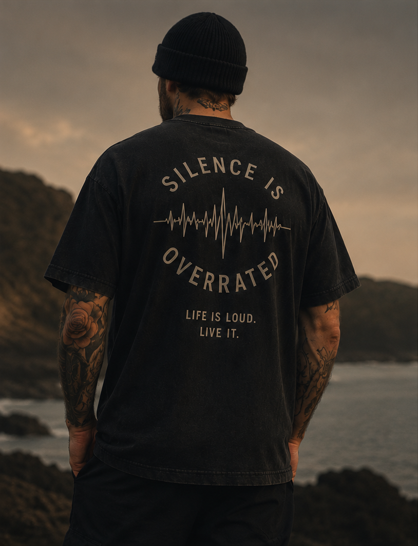 Back view of a tattooed man in a black Tinnitus Gang tee printed with "Silence Is Overrated" by the coast at dusk